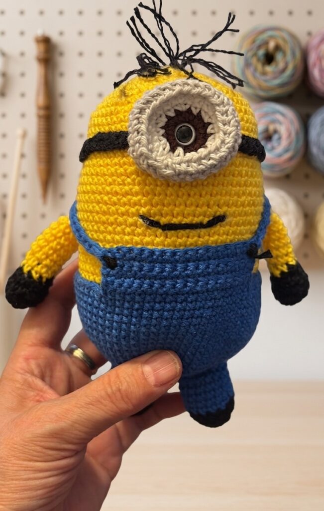 Amigurumi de Minion tejido a crochet, sostenido con la mano, con peto azul, un ojo y detalles negros.