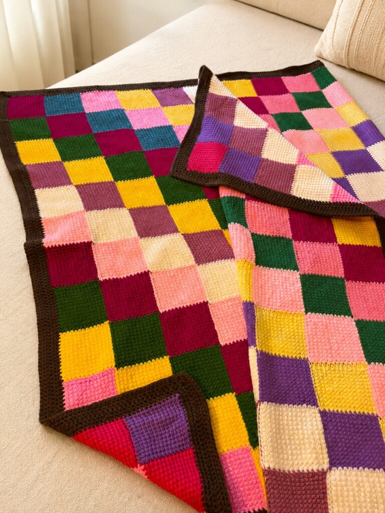 Manta tejida en punto tunecino con cuadrados de colores y borde marrón colocada sobre un sofá claro.